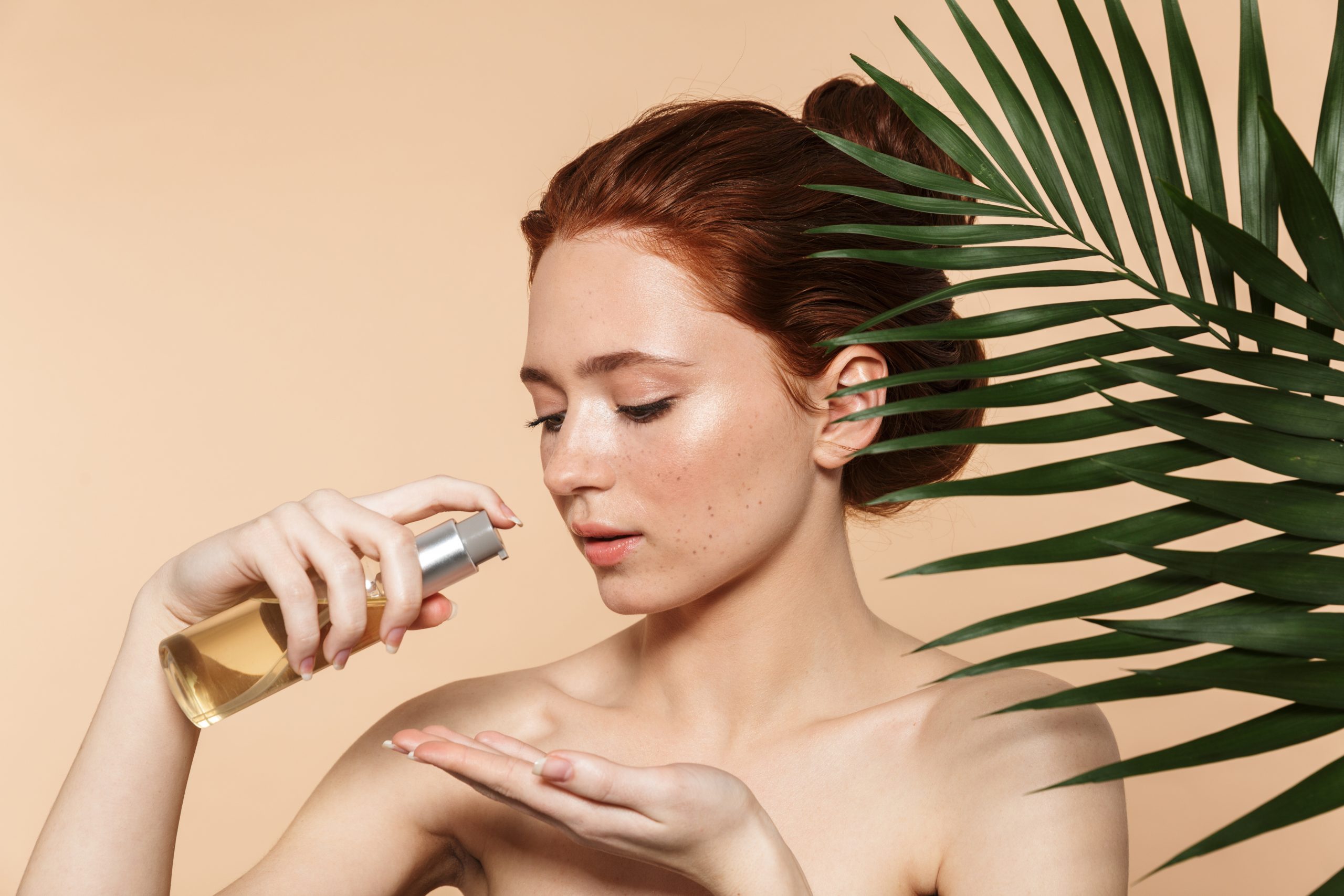 Beauty portrait of an attractive young topless redhead woman standing isolated over beige background, applying body oil, posing surrounded with tropical leaves